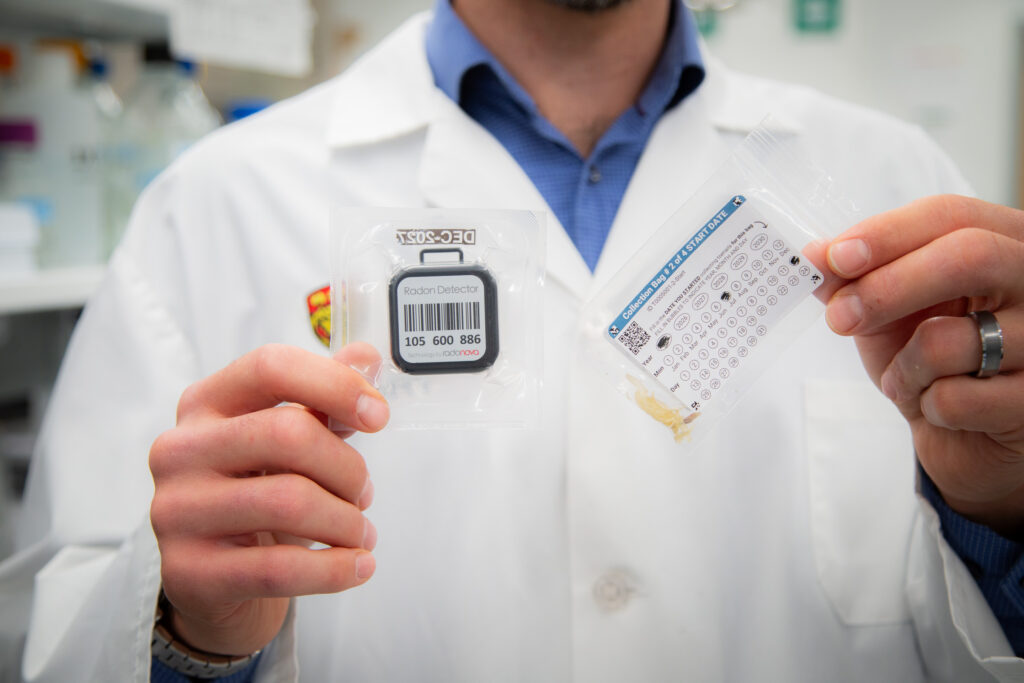 Dr. Dustin Pearson holding a radon test kit and a toenail sample collection bag (Credit: Glynnis Mutch Photography)